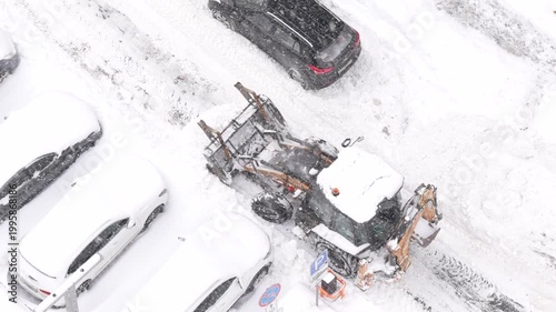 Snowplow tractor clearing snow between parked cars in residential area, winter urban maintenance work