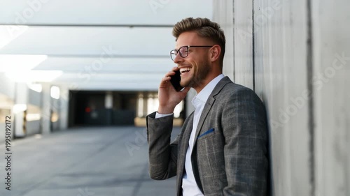 Caucasian male young businessman in grey suit and glasses leaning on wall smiling while on smartphone, relaxed urban executive portrait with contemporary architecture setting