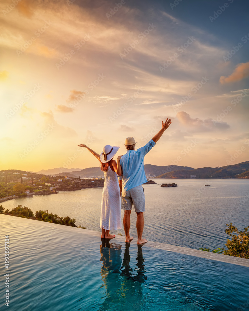 Fototapeta premium A happy couple on summer vacation standing by the swimming pool enjoys a beautiful sunset behind the sea