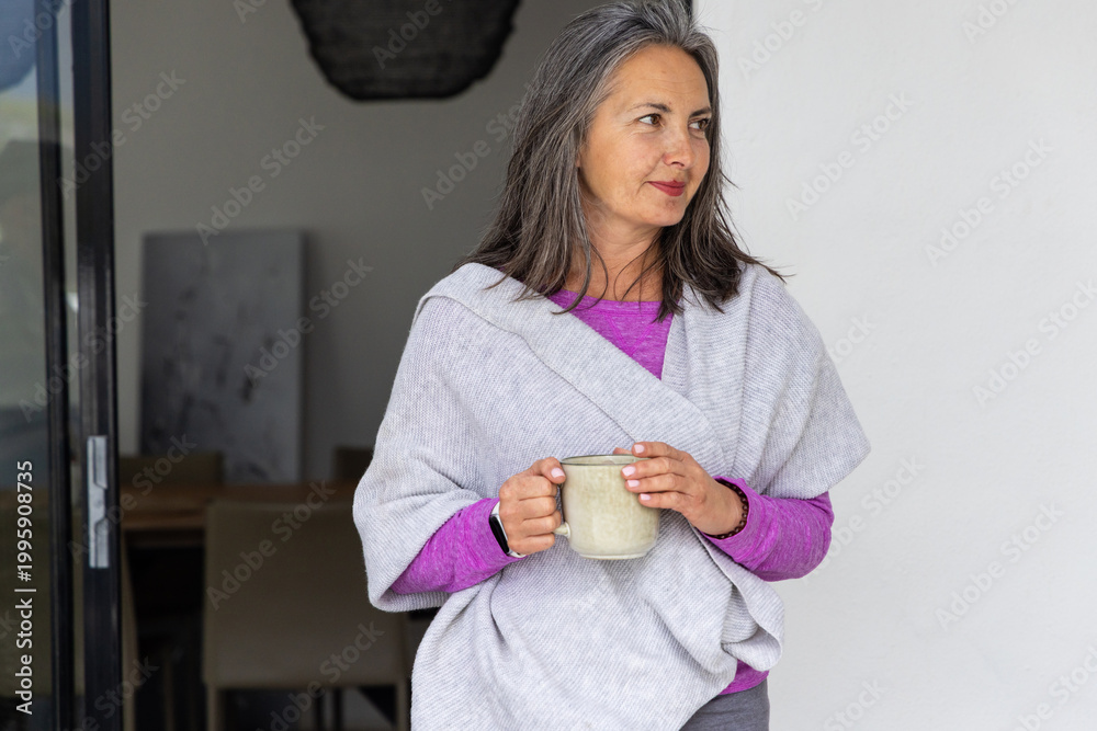 Naklejka premium Senior woman standing near sliding glass door holding ceramic mug, wearing magenta top and wrap