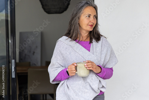 Senior woman standing near sliding glass door holding ceramic mug, wearing magenta top and wrap