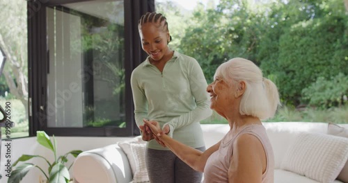 Adult African American caregiver gently taking Senior woman's hand, guiding arm exercise on sofa