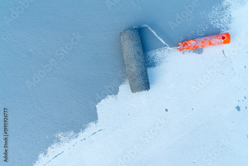 Paint roller with orange handle applying blue paint on a textured white concrete surface. Renovation, home improvement, and construction concept showing professional wall painting progress.