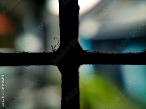 Close-up of a window grille, showing intersecting wooden bars with visible dust and cobwebs. The background is blurred, displaying a mix of colors, suggesting an outdoor set