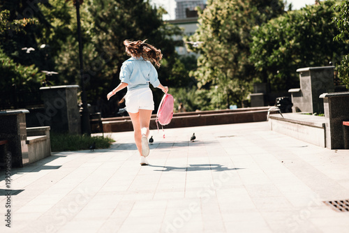 Obraz na płótnie Young woman in casual summer outfit runs through a sunny park in the city carryi