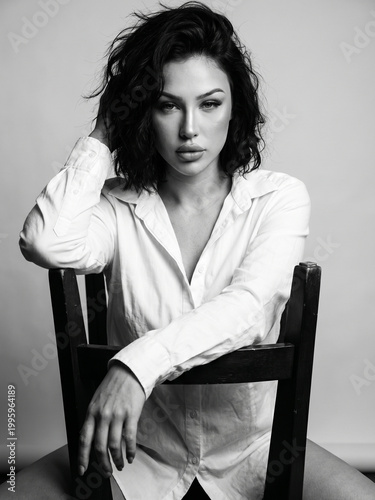 Dramatic black and white portrait of a beautiful young woman with messy bob hair sitting on a wooden chair, moody studio lighting