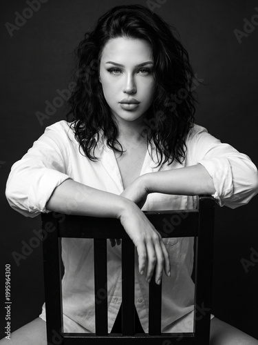 Dramatic black and white portrait of a beautiful young woman with messy bob hair sitting on a wooden chair, moody studio lighting