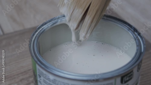Close-up of a gloved hand dipping a paintbrush into a can of white paint on a wooden surface. Home renovation, DIY painting, wood treatment, and repair concept with copy space.