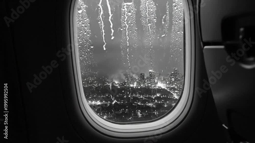 Rain drops stream down airplane window with nighttime city lights in black and white during flight