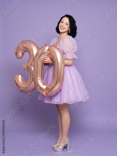 Happy young woman celebrating 30th birthday holding rose gold number balloons, studio portrait on purple background