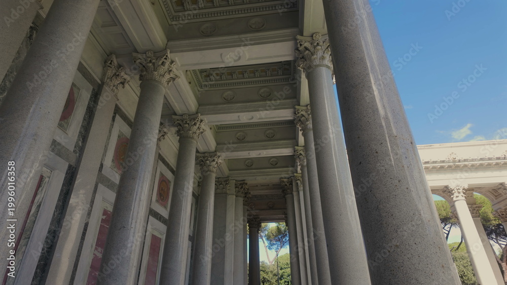 Fototapeta premium Row of tall stone columns with ornate capitals supporting decorated ceiling in classical architecture, exterior view with blue sky and trees in the background