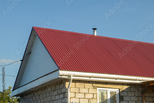 Red metal roof with vertical ribbing on a stone house under clear blue sky, chimney visible on the roofline, showcasing modern architectural design elements