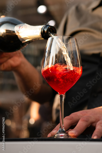 Close-up of a bartender pouring sparkling wine into a glass with a vivid red cocktail, capturing bubbles and motion in warm evening light.