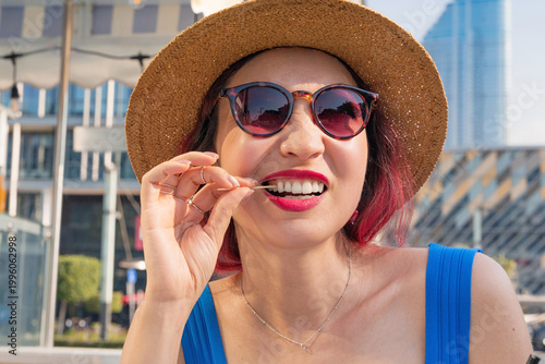 Happy woman wearing sunglasses and a straw hat while using a toothpick, celebrating vacation lifestyle in the urban landscape
