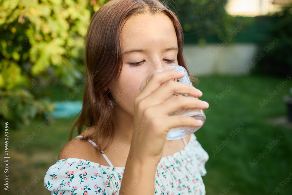 Fototapeta premium Girl drinks clear water from glass in sunny garden while enjoying nature in the afternoon light
