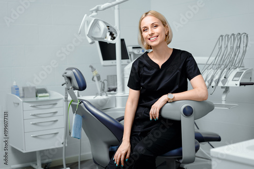 Young blonde woman dentist posing at modern dental cabinet next to dental chair cheerfully smiling at camera. Pretty stomatologist at dental clinic