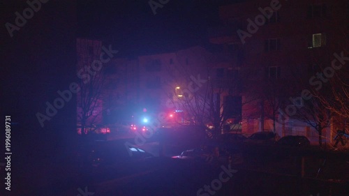 Night street illuminated by flashing lights, emergency vehicles respond outside apartment building, red and blue strobes cast silhouettes and shadows, siren wail and urgent