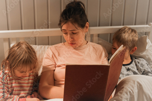 Family bedtime stories. A mother reads a story to her children, wrapped in a blanket. Children listen to soothing stories. A cozy family scene before bed in a softly lit bedroom. Copy space. Banner