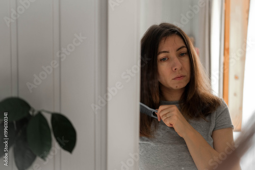 A woman combs her hair in front of a mirror. She's tidying herself up. She's wearing a casual gray T-shirt. Her expression is focused, as she prepares for the workday. Self-care. Space for text.