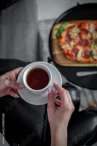 Female hands holding a white cup of tea in the foreground with pizza on a wooden board blurred in the background indoors