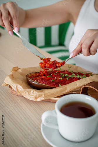 Woman eating baked stuffed eggplant with rich tomato sauce at wooden table, cozy café serving, close lifestyle food scene with hot tea
