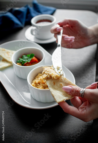 Female hands preparing flatbread with hummus from a white appetizer platter with roasted vegetable dip and tea on a dark table