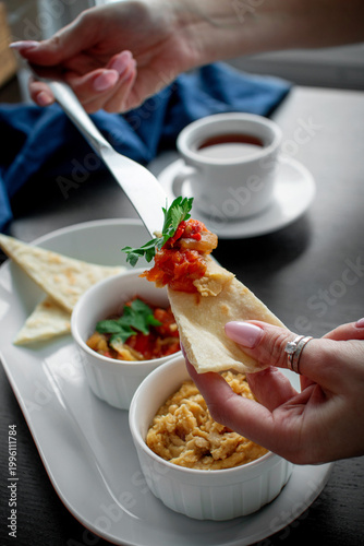 Female hands topping flatbread with roasted vegetable dip and hummus from a white appetizer platter with tea on a dark table