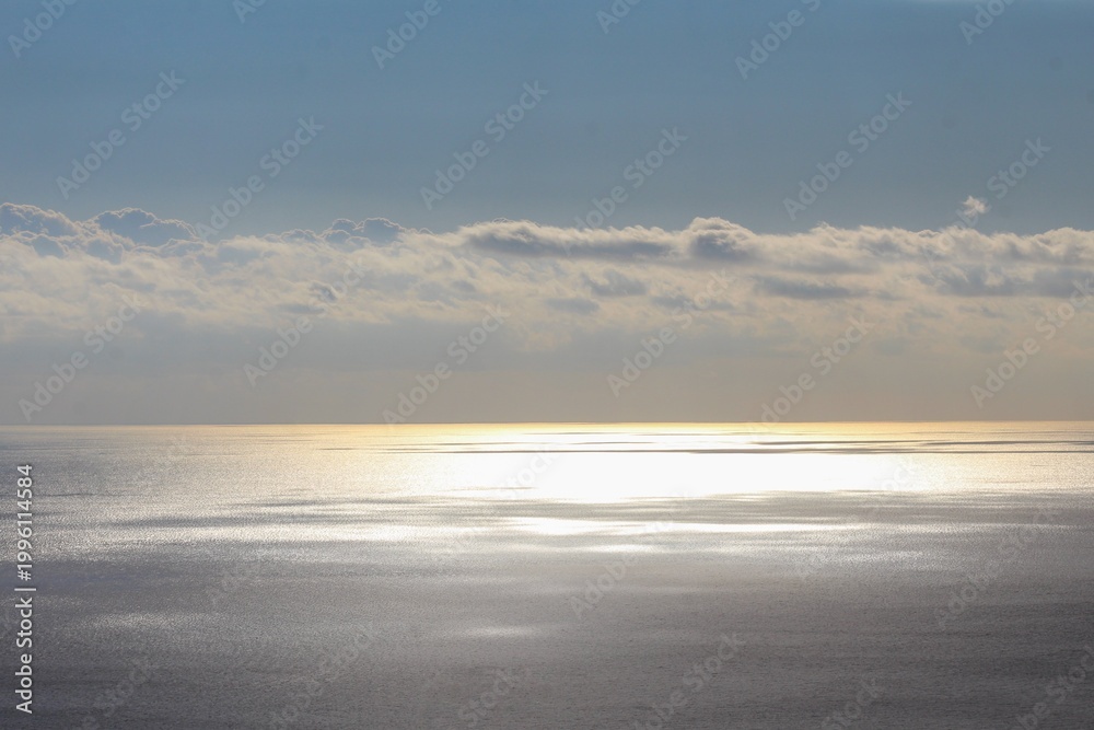 Obraz premium A tranquil seascape where sunlight pierces through clouds and glistens across the calm water surface. The soft golden reflections and layered clouds create a peaceful, atmospheric horizon scene.