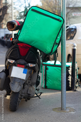 Vertical street city defocused photo. Back view. Green thermal bag for delivery food, drink without logo on black moped baggage rack on parking lot. Concept of dinner, lunch