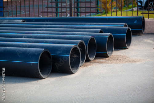 Stack of new black HDPE plastic pipes with blue stripes for cold water supply. Industrial construction site and municipal infrastructure background