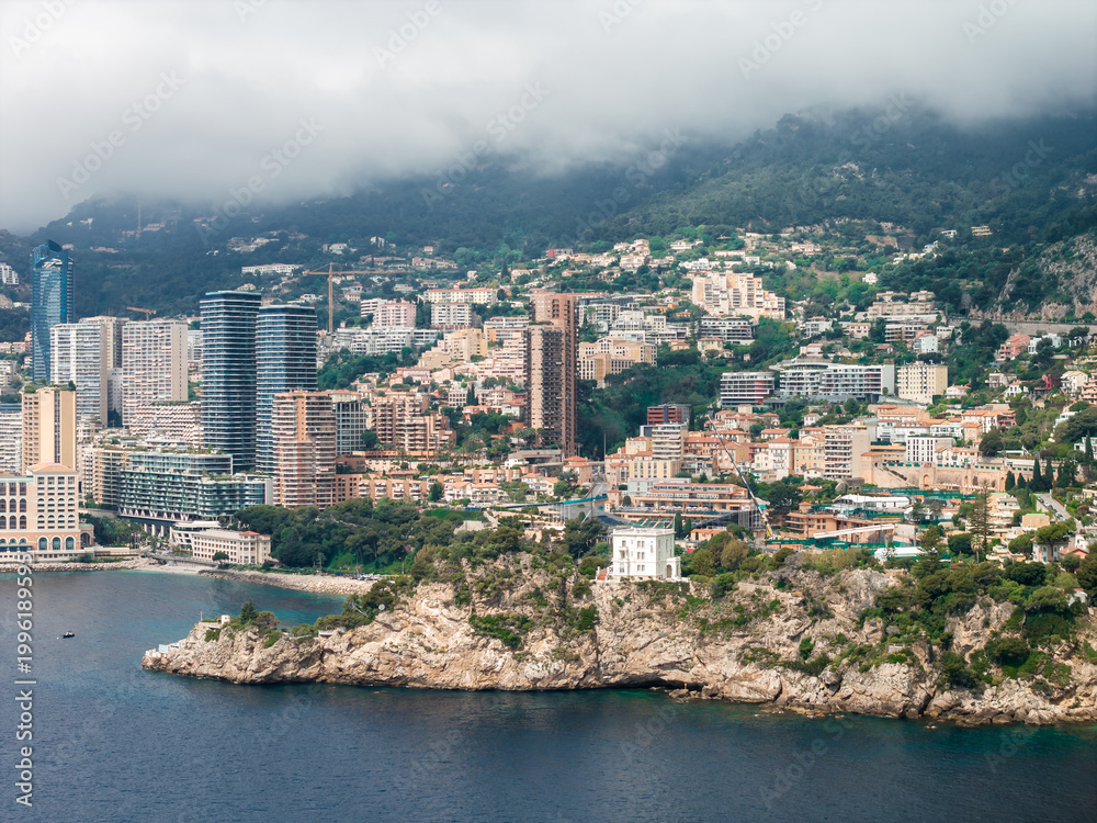 Fototapeta premium Aerial view of Monaco on the French Riviera shows towers and terraced apartments on steep hills, a curved bay, pale cliffs, a white villa, and turquoise water under overcast skies.