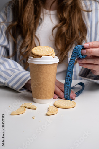 Woman holding coffee cup with cookies and blue measuring tape on table. Dietary control concept. 