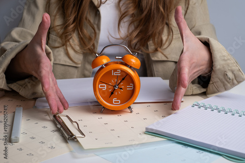 Woman’s hands framing orange alarm clock on desk with calendars and notebooks. Urgency and time management concept.