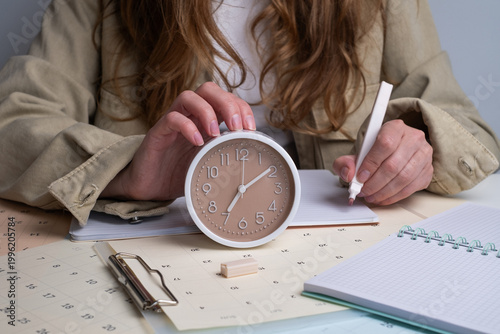 Woman holding alarm clock and writing in notebook with calendar on desk. Effective time management concept. 
