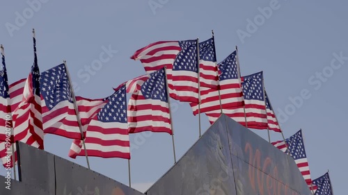 A majestic, slow-motion video of multiple United States national flags lined along the top of a building. The flags flutter gracefully in the wind against a clear sky.