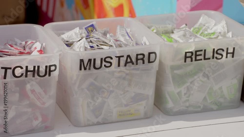 This close up, panning video shows ketchup, mustard, and relish condiments in containers at an outdoor vendor's stand. 