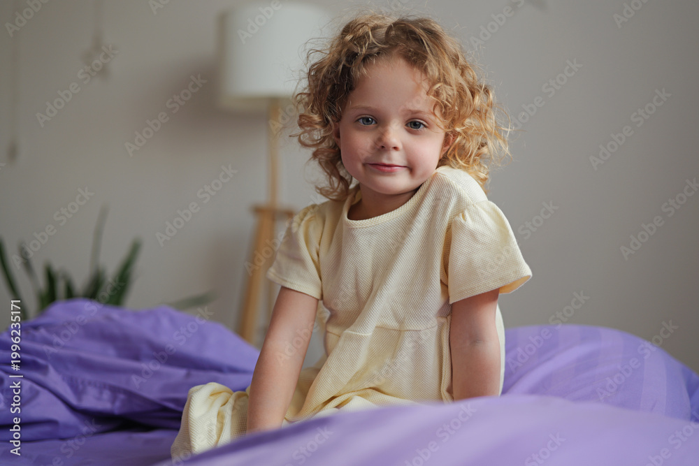 Fototapeta premium A child with curly hair is sitting on a purple duvet cover in a bright room. She wears a yellow dress and looks toward the camera while the sun shines in