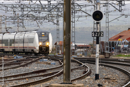 Commuters and travellers gather along the platforms at Ávila railway station as a regional train arrives, framed by overhead cables and urban architecture