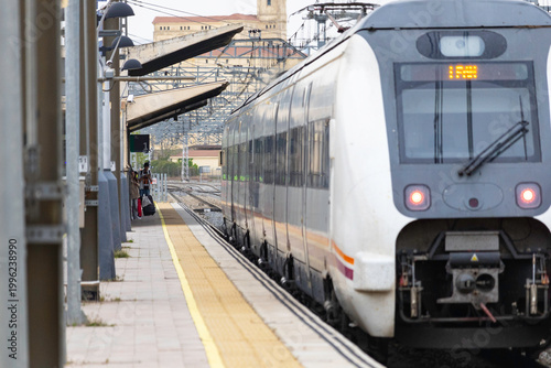 Commuters and travellers gather along the platforms at Ávila railway station as a regional train arrives, framed by overhead cables and urban architecture