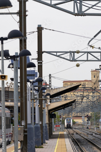 Commuters and travellers gather along the platforms at Ávila railway station as a regional train arrives, framed by overhead cables and urban architecture