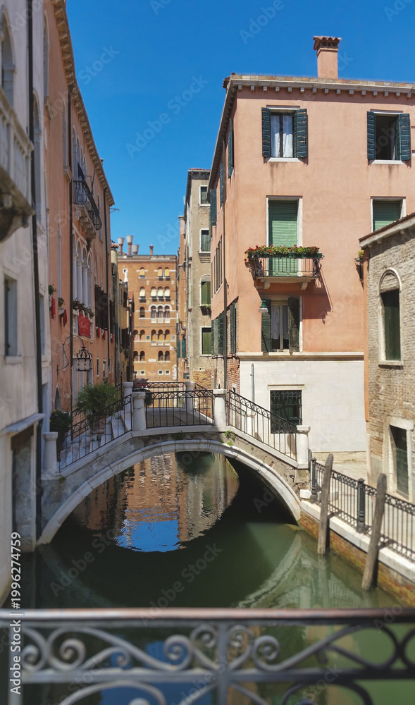 Naklejka premium Scenic view of a small arched stone bridge crossing a narrow canal with historic Venetian houses