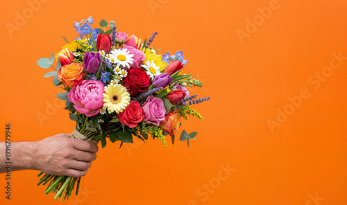 Hand holding a vibrant mixed dopamine bouquet of colorful flowers against a bold orange background, featuring roses, tulips, and daisies in a bright, clean studio composition.