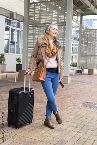 Walking woman wearing tan jacket pulling black suitcase at entry, with crossbody bag and phone