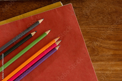 Seven pencils resting diagonally on red notebook with yellow edge on left wooden desk, copy space