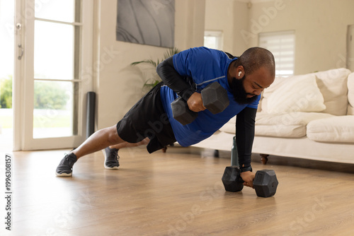 African American man doing renegade row at home wearing workout gear with hex dumbbells and earbuds