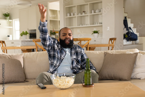 African American middle-aged man shouting and gesturing while sitting on sofa with popcorn, remote