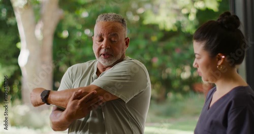 Senior African American man feeling shoulder pain doing arm stretch on patio while Asian PT guiding