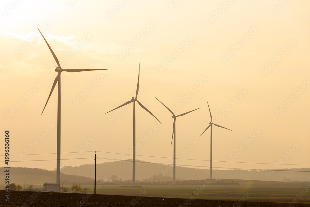 Fototapeta premium Wind turbines stand in a field under a golden sky during sunset in a rural area