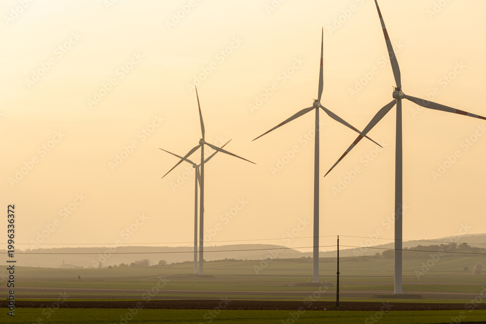 Fototapeta premium Wind turbines stand tall in a field at sunset near a rural area