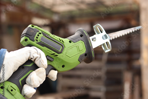 A craftsman in a protective glove holds a compact green cordless reciprocating saw. The tool is designed for fast and efficient sawing of wood, metal, plastic and plywood. Power tool.